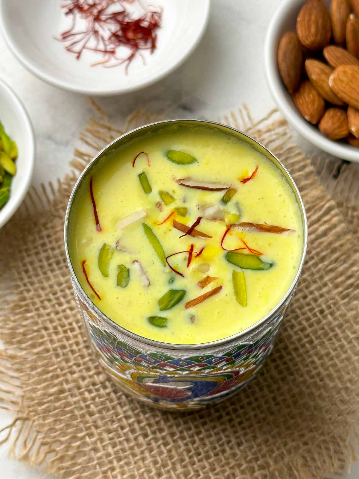 badam milk (badam doodh) served in a steel glass garnished with nuts and saffron with nits on the side