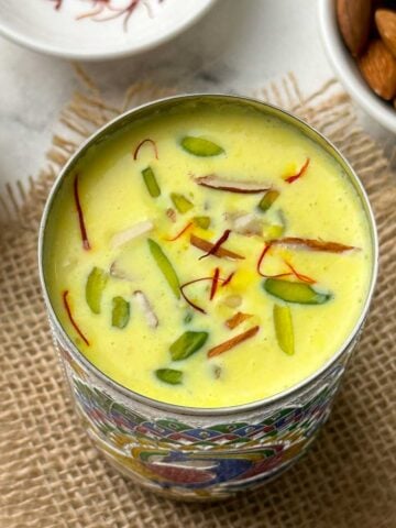 badam milk served in a steel glass garnished with nuts and saffron with nits on the side