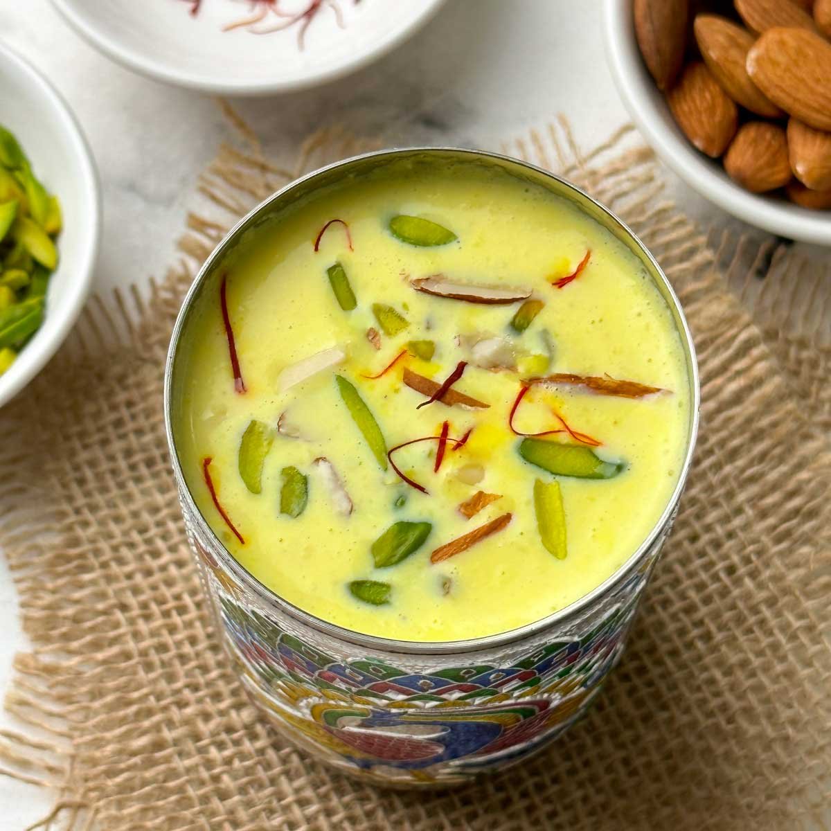 badam milk served in a steel glass garnished with nuts and saffron with nits on the side