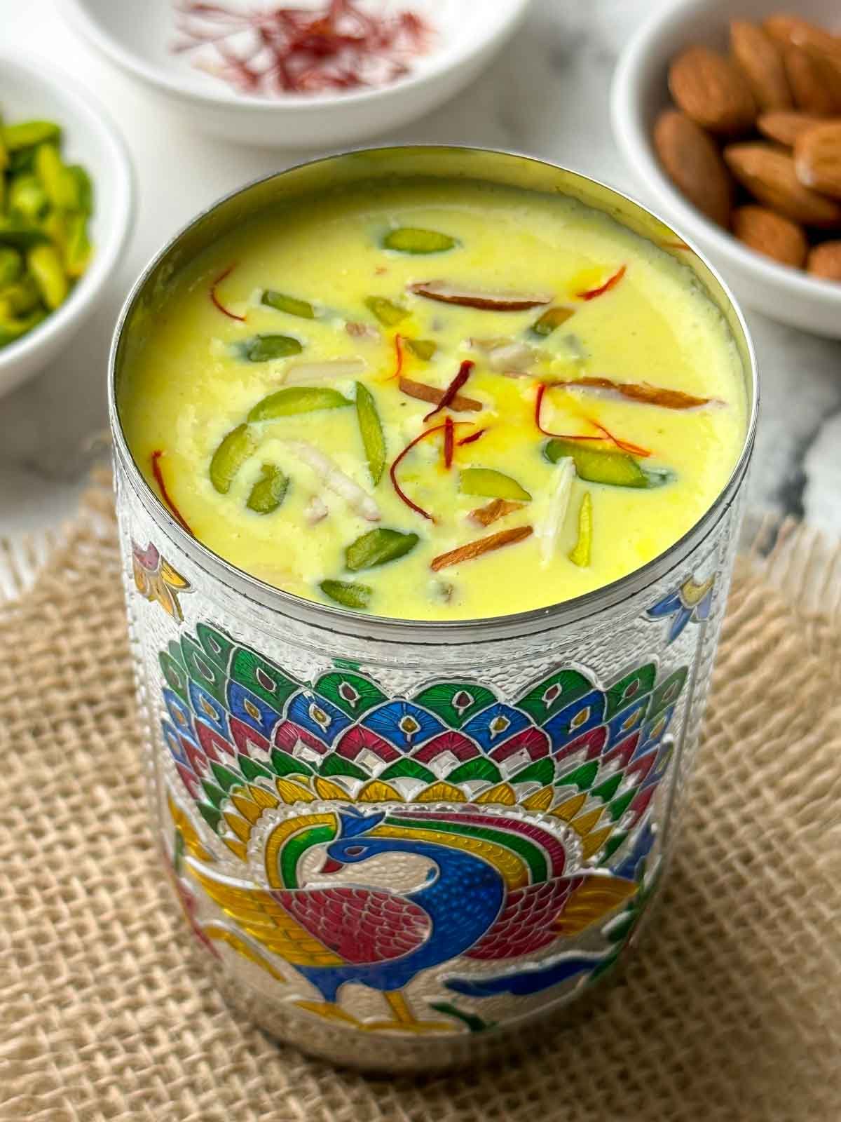 badam milk served in a steel glass garnished with nuts and saffron with nits on the side