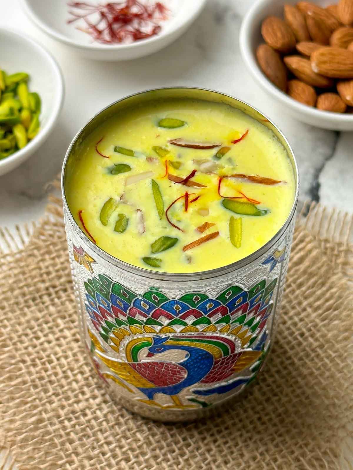 hot badam milk served in a steel glass garnished with nuts and saffron with nits on the side