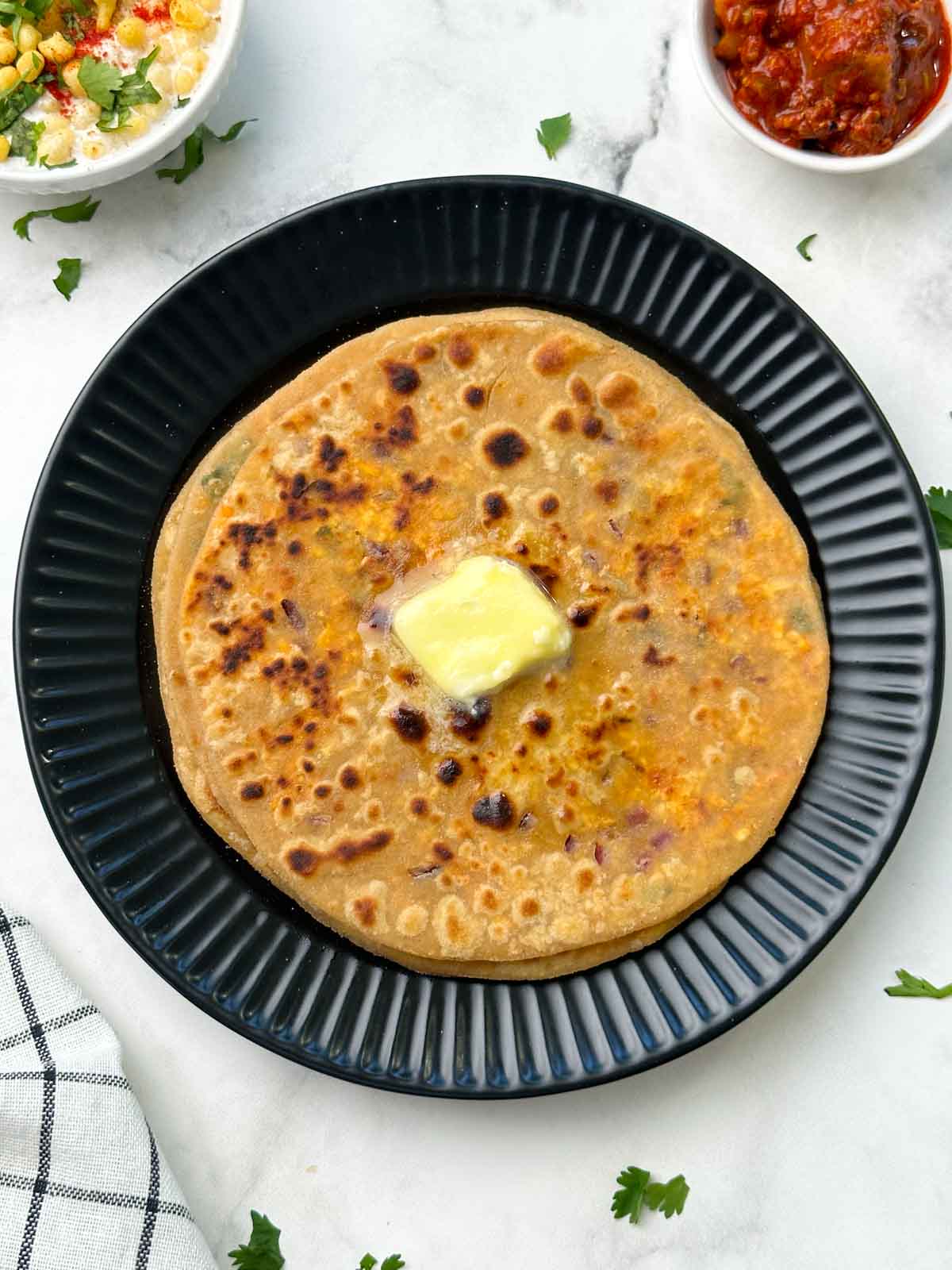 stack of paneer parathas on a black plate with butter on top and raita and pickle on the side