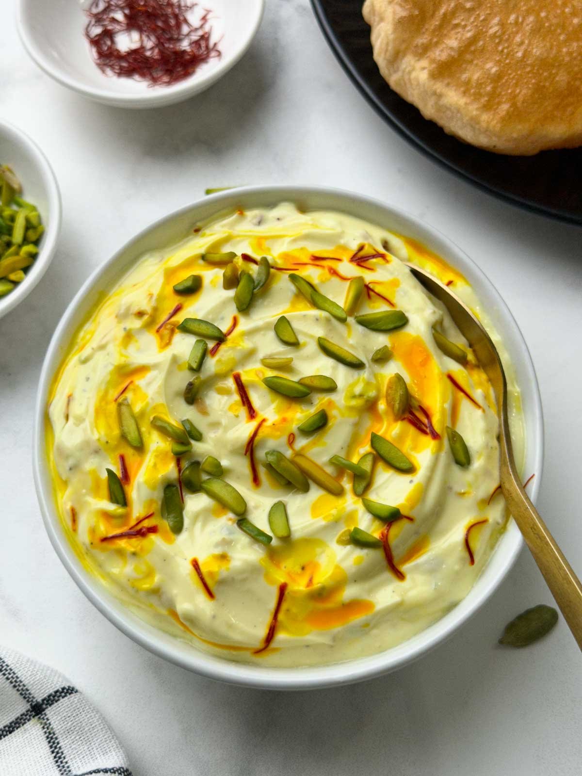 shrikhand served in a bowl with a spoon with poori, pista on the side