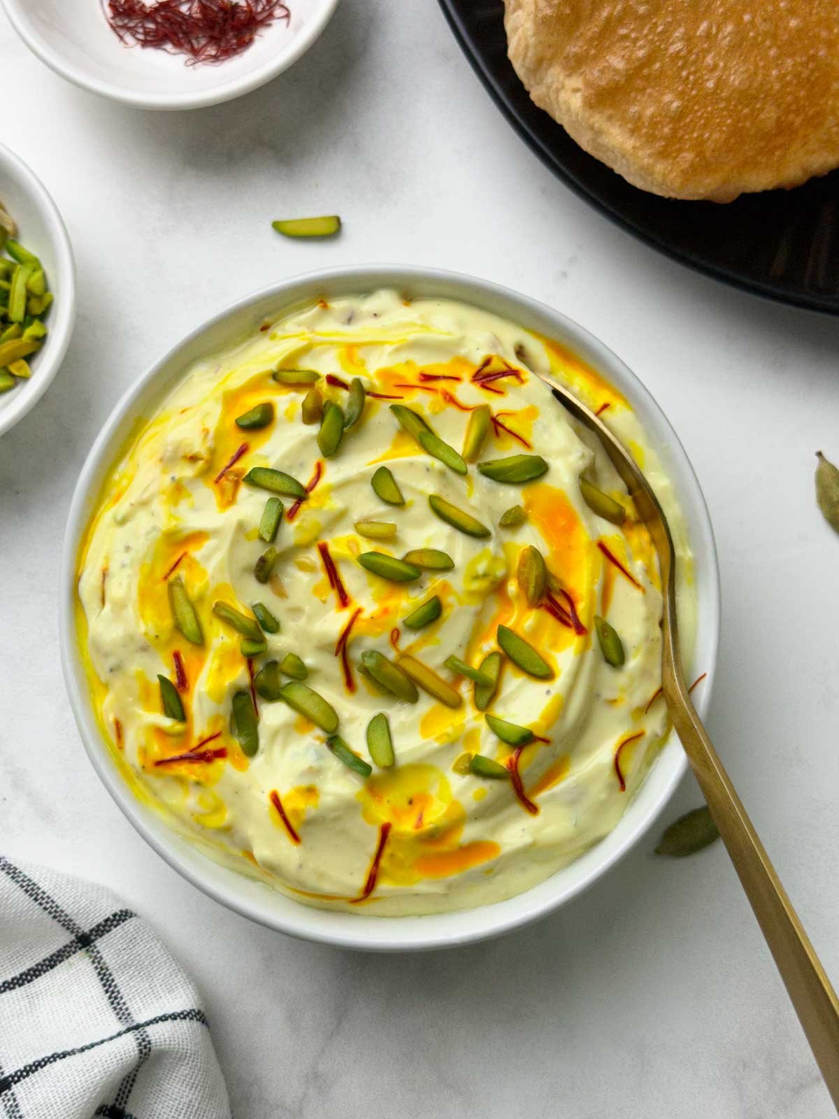 shrikhand served in a bowl with a spoon with poori, pista on the side