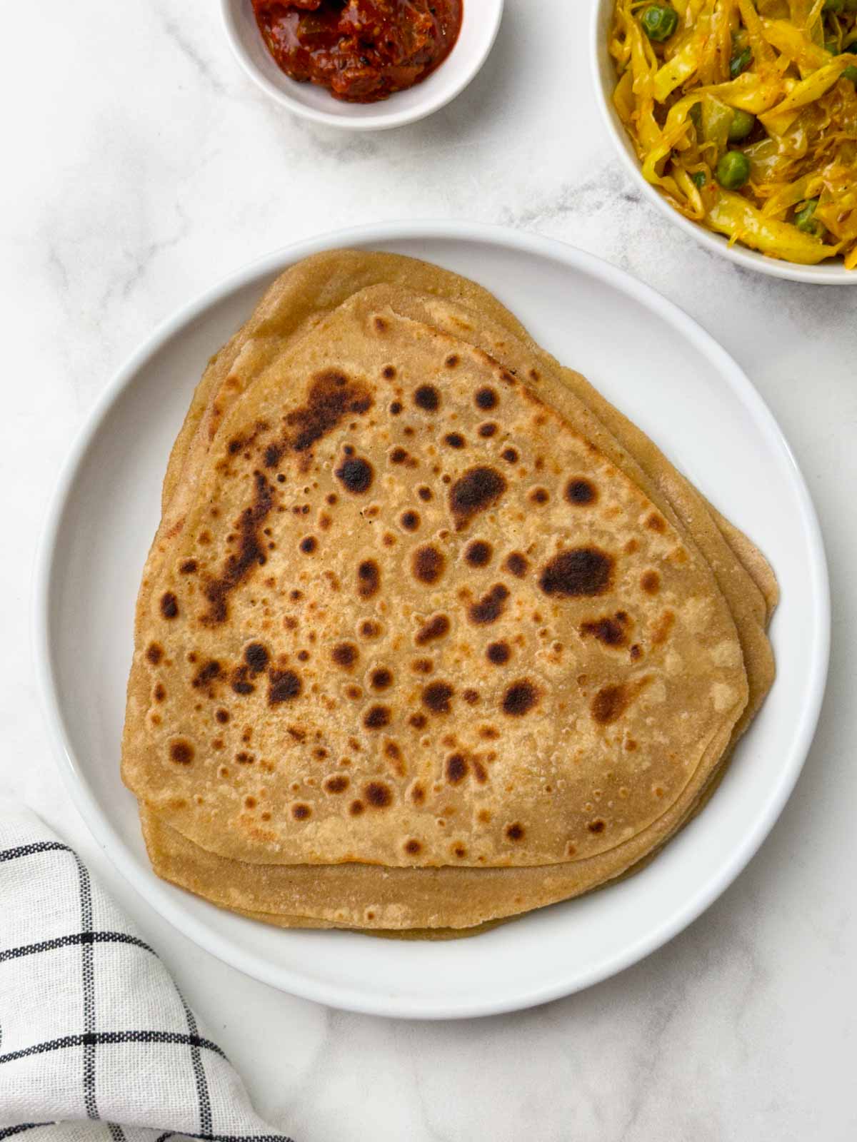 Stack of triangular shape paratha with cabbage stir fry and pickle on the side.