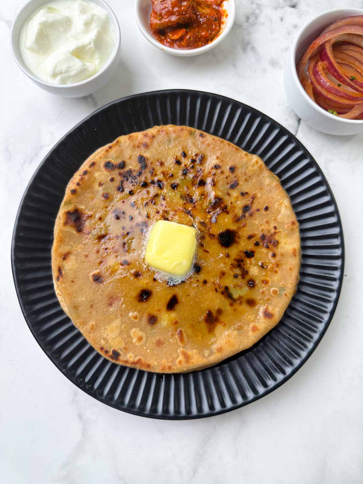 aloo paratha stacked on a plate with butter on the top with yogurt, laccha onion on the side