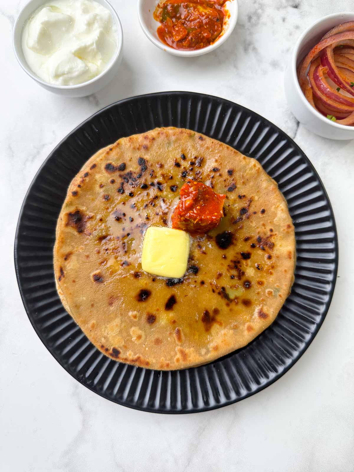 aloo paratha stacked on a plate with butter and pickle on the top with yogurt, laccha onion on the side