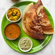 Two crispy masala dosa served on a banana leaf plastic plate along with potato masala, coconut chutney and vegetable sambar on the side and filter coffee in the cups