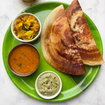 Two crispy masala dosa served on a banana leaf plastic plate along with potato masala, coconut chutney and vegetable sambar on the side and filter coffee in the cups