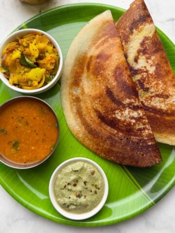 Two crispy masala dosa served on a banana leaf plastic plate along with potato masala, coconut chutney and vegetable sambar on the side and filter coffee in the cups