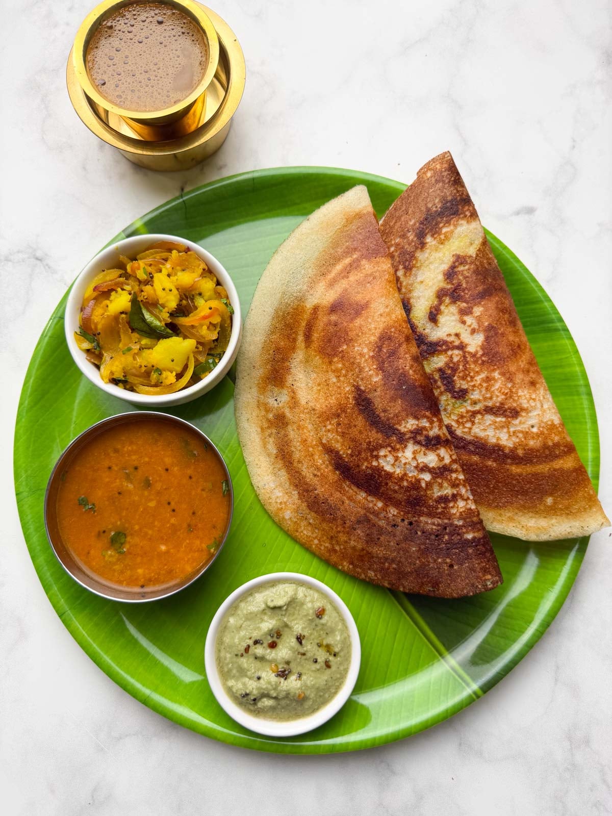 Two crispy masala dosa served on a banana leaf plastic plate along with potato masala, coconut chutney and vegetable sambar on the side and filter coffee in the cups