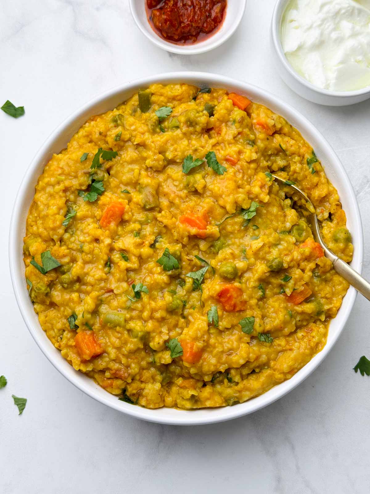vegetable dalia khichdi (broken wheat khichdi) served in a bowl with a spoon and pickle and yogurt on the side