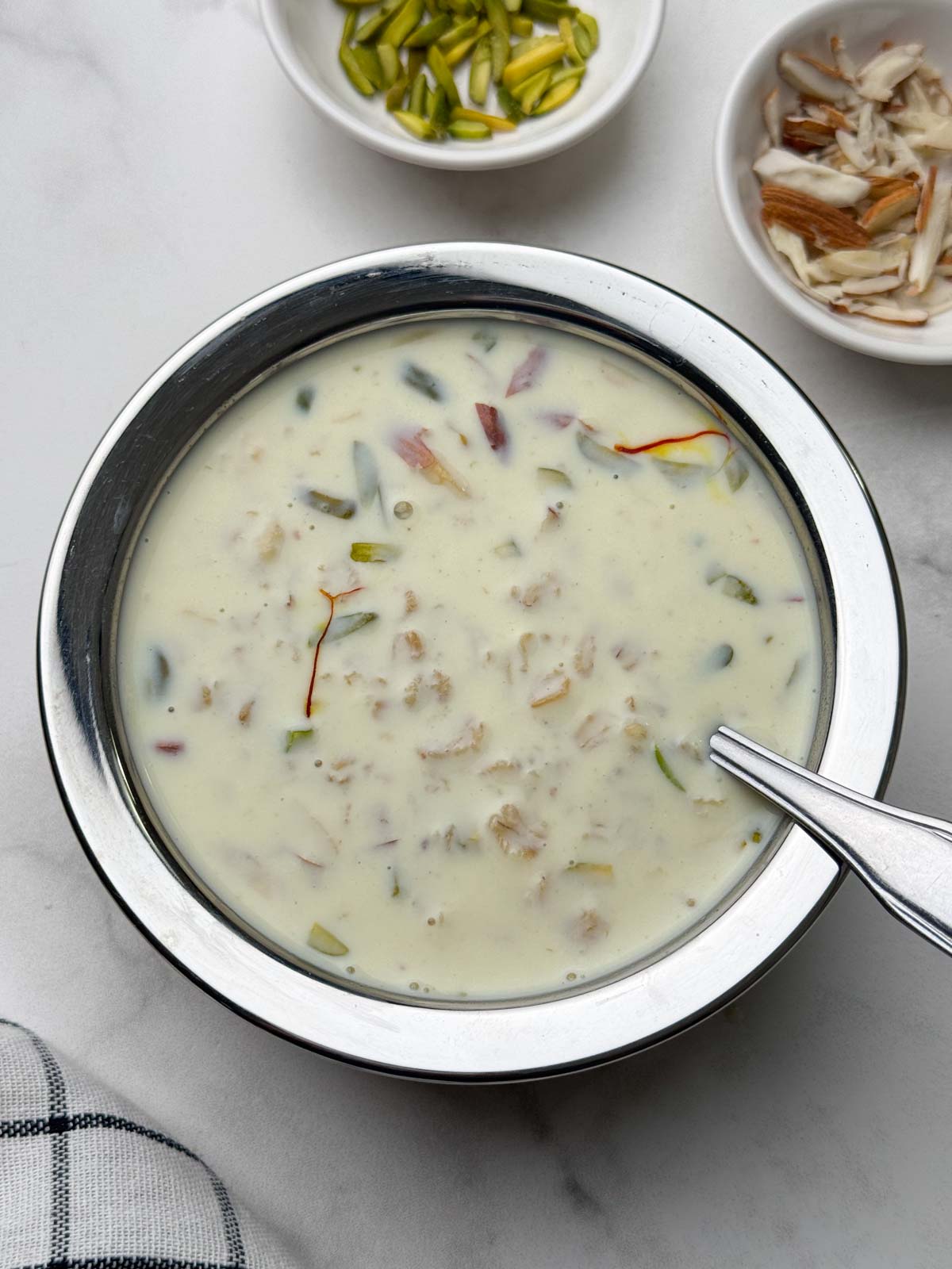 oats kheer served in a steel bowl with a spoon and nuts on the side