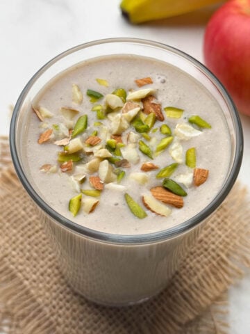 apple banana smoothie in a glass garnished with nuts with fruit in the background