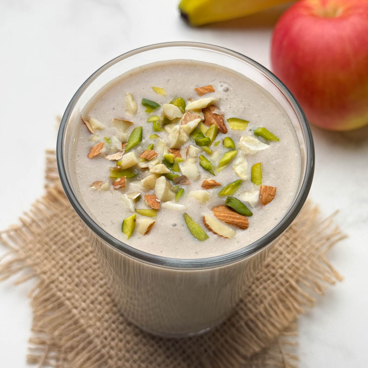 apple banana smoothie in a glass garnished with nuts with fruit in the background