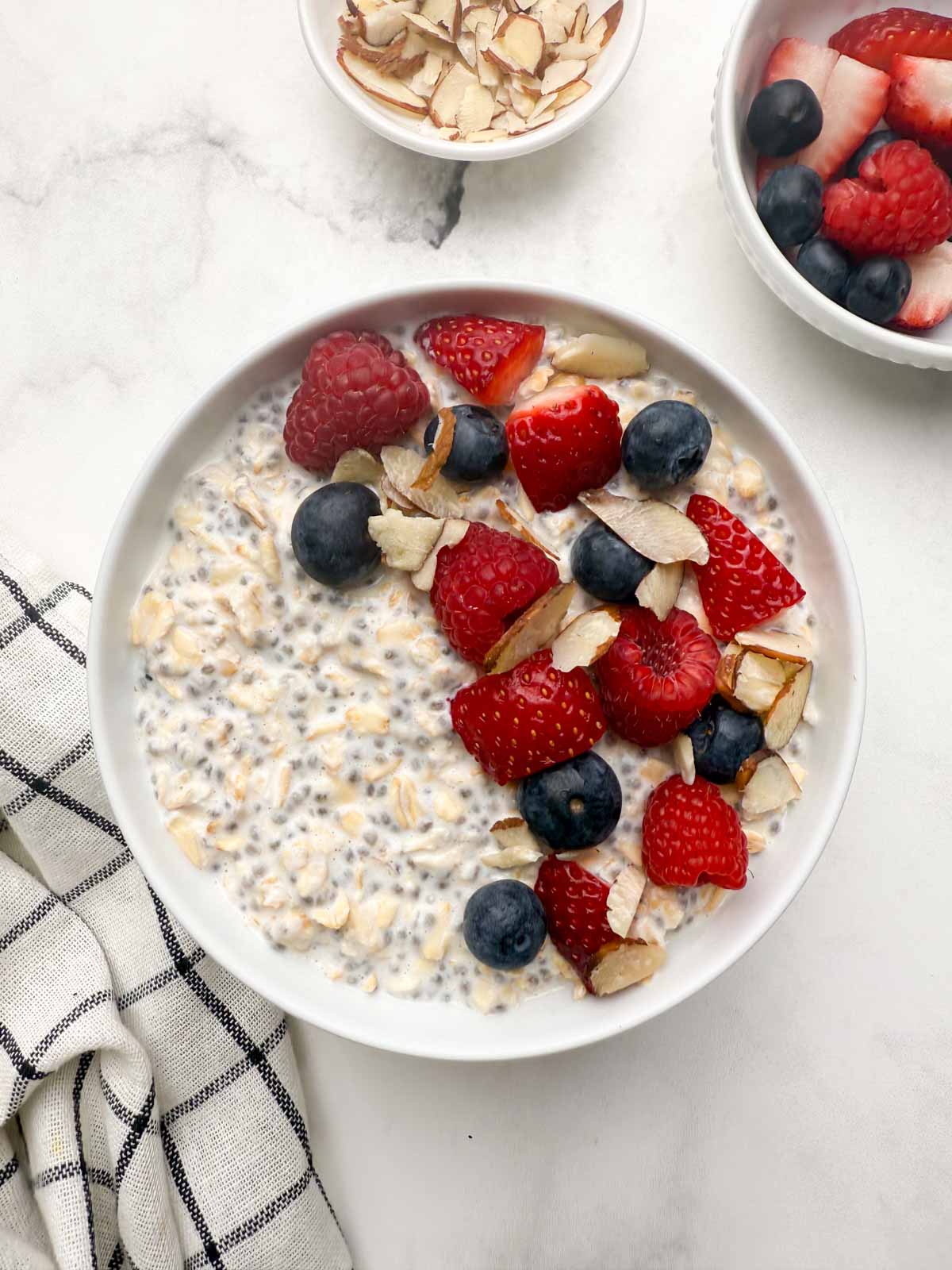 Overnight oats with chia seeds in a white serving bowl with berries and slivered almonds on the top and with two bowl each with berries and almonds in the side.
