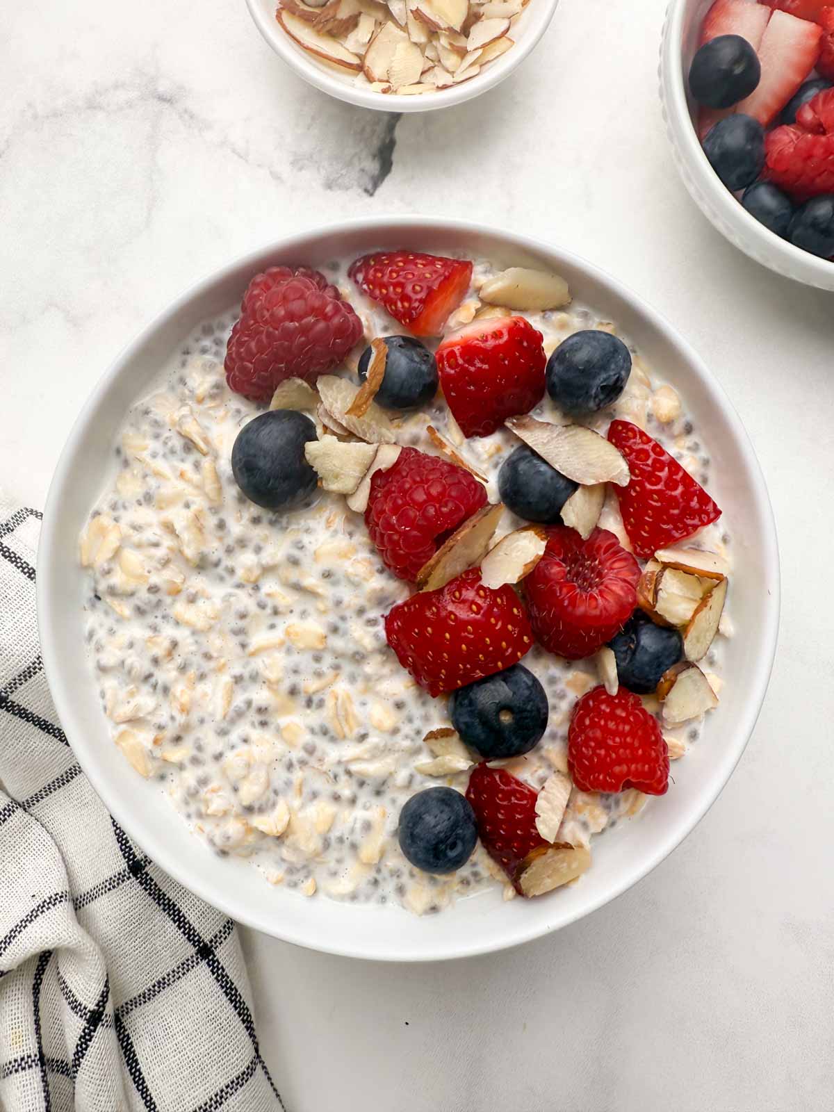 Overnight oats with chia seeds in a white serving bowl with berries and slivered almonds on the top and with two bowl each with berries and almonds in the side.