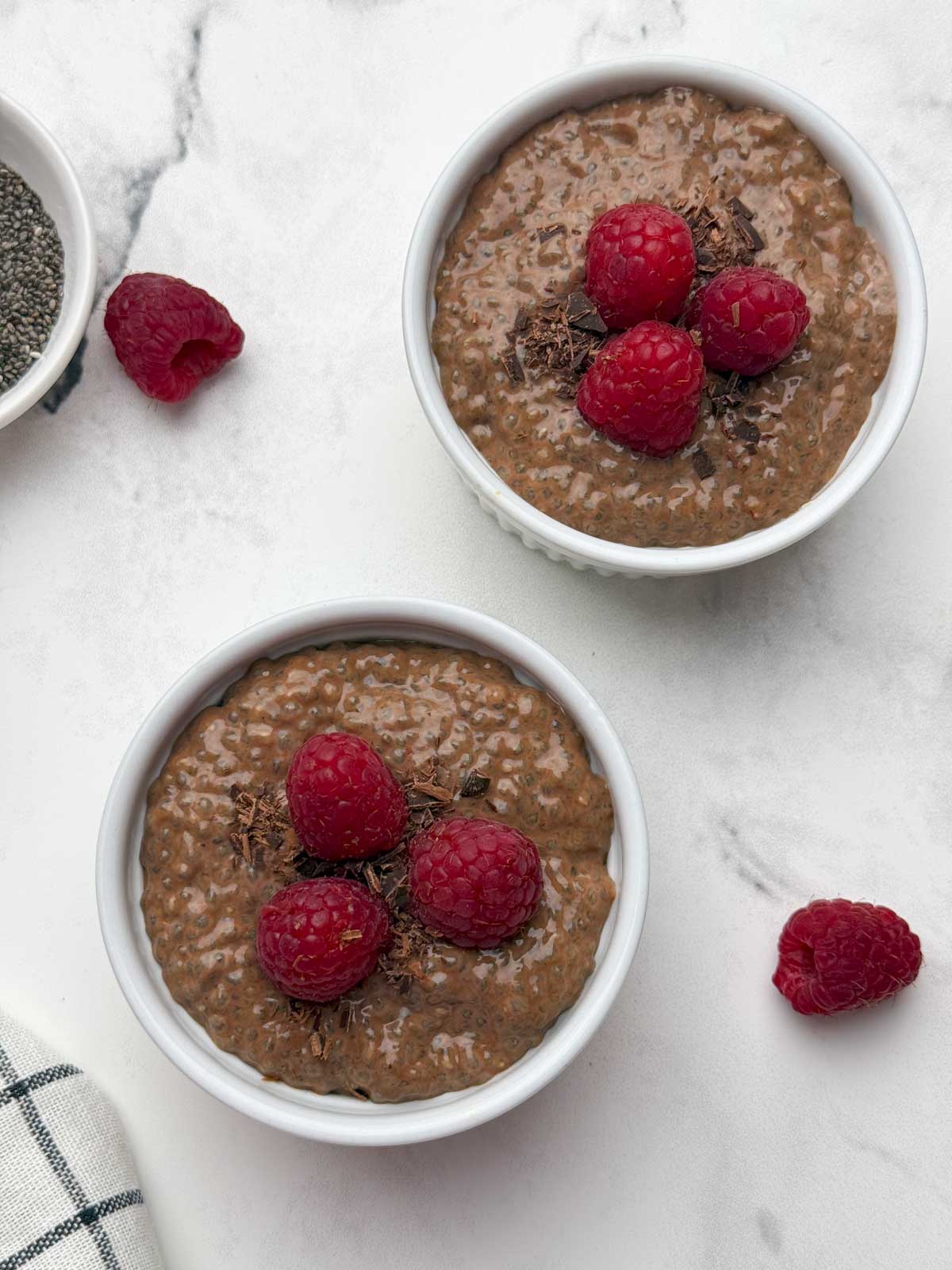 Two ramkens filled with chocolate chia pudding topped with raspberries and chocolate shavings and a bowl of raw chia seeds and raspberries on the side.