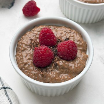 Two ramkens filled with chocolate chia pudding topped with raspberries and chocolate shavings and a bowl of raw chia seeds on the side.
