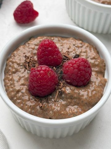 Two ramkens filled with chocolate chia pudding topped with raspberries and chocolate shavings and a bowl of raw chia seeds on the side.