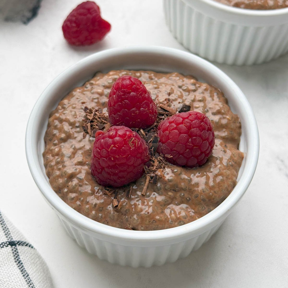 Two ramkens filled with chocolate chia pudding topped with raspberries and chocolate shavings and a bowl of raw chia seeds on the side.