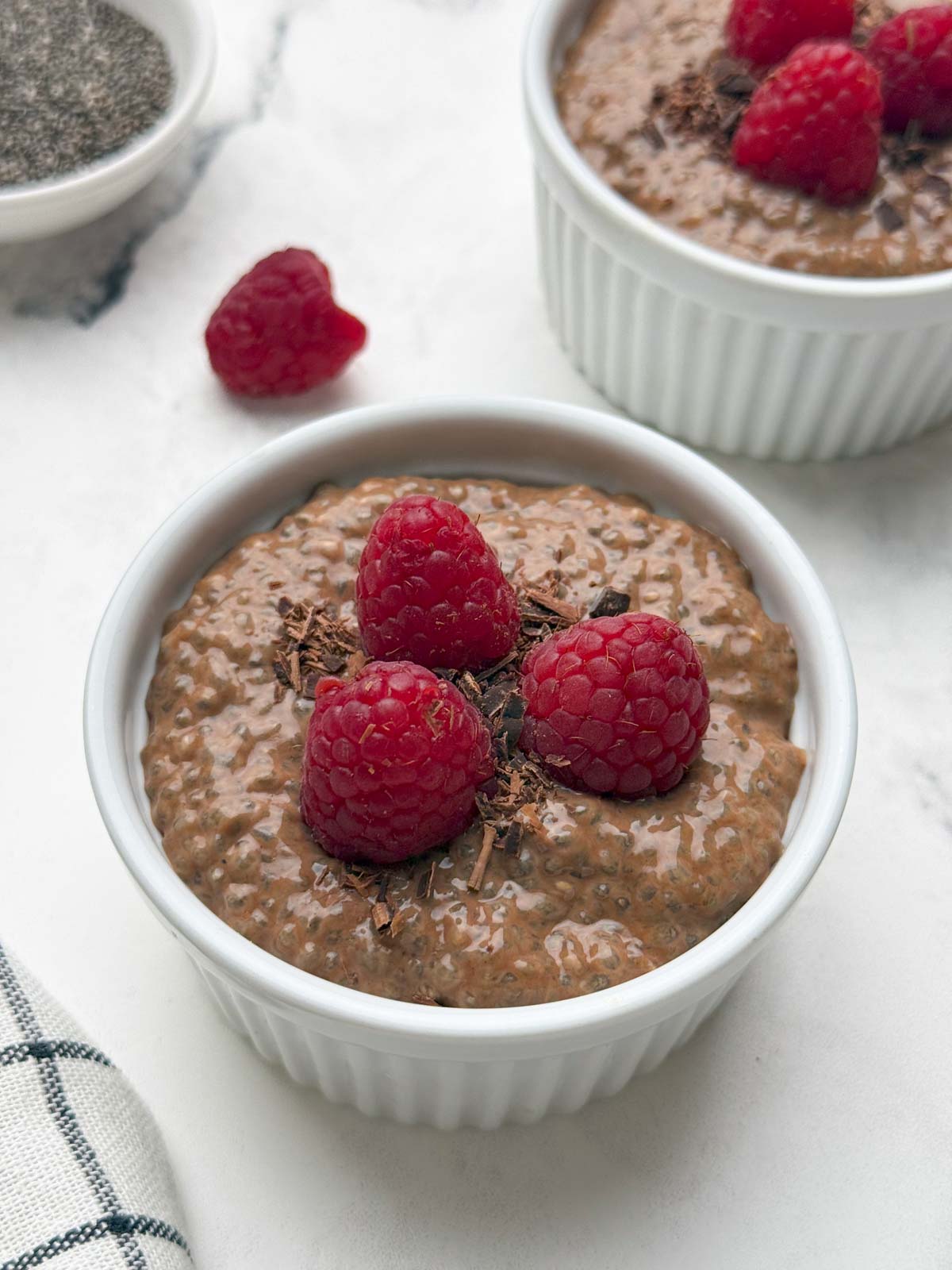 Two ramkens filled with chocolate chia pudding topped with raspberries and chocolate shavings and a bowl of raw chia seeds on the side.