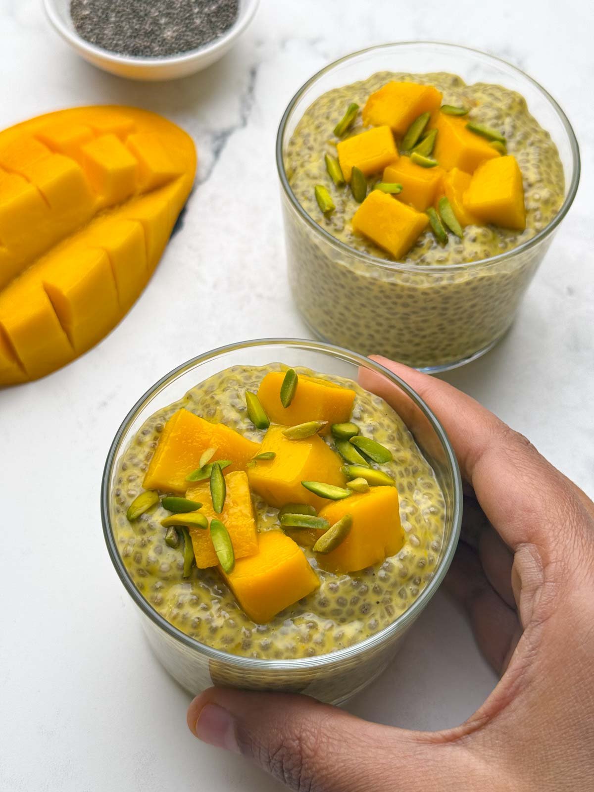 Holding one glass bowl filled with mango chia pudding topped with diced mangoes and pista. You can also see half cut mango and another serving bowl with chia pudding on the side.