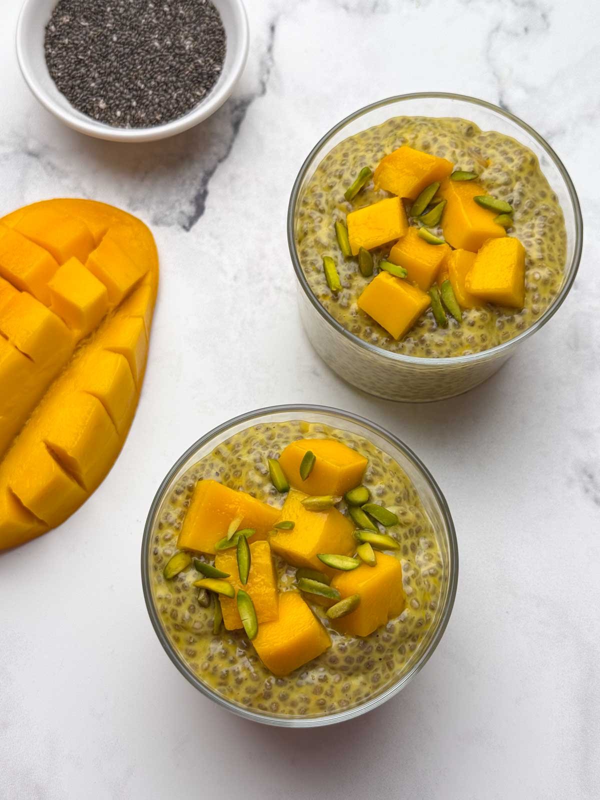Two serving bowl with mango chia pudding topped with diced mangoes and pista. You can also see half cut mango and chia seeds in a bowl.