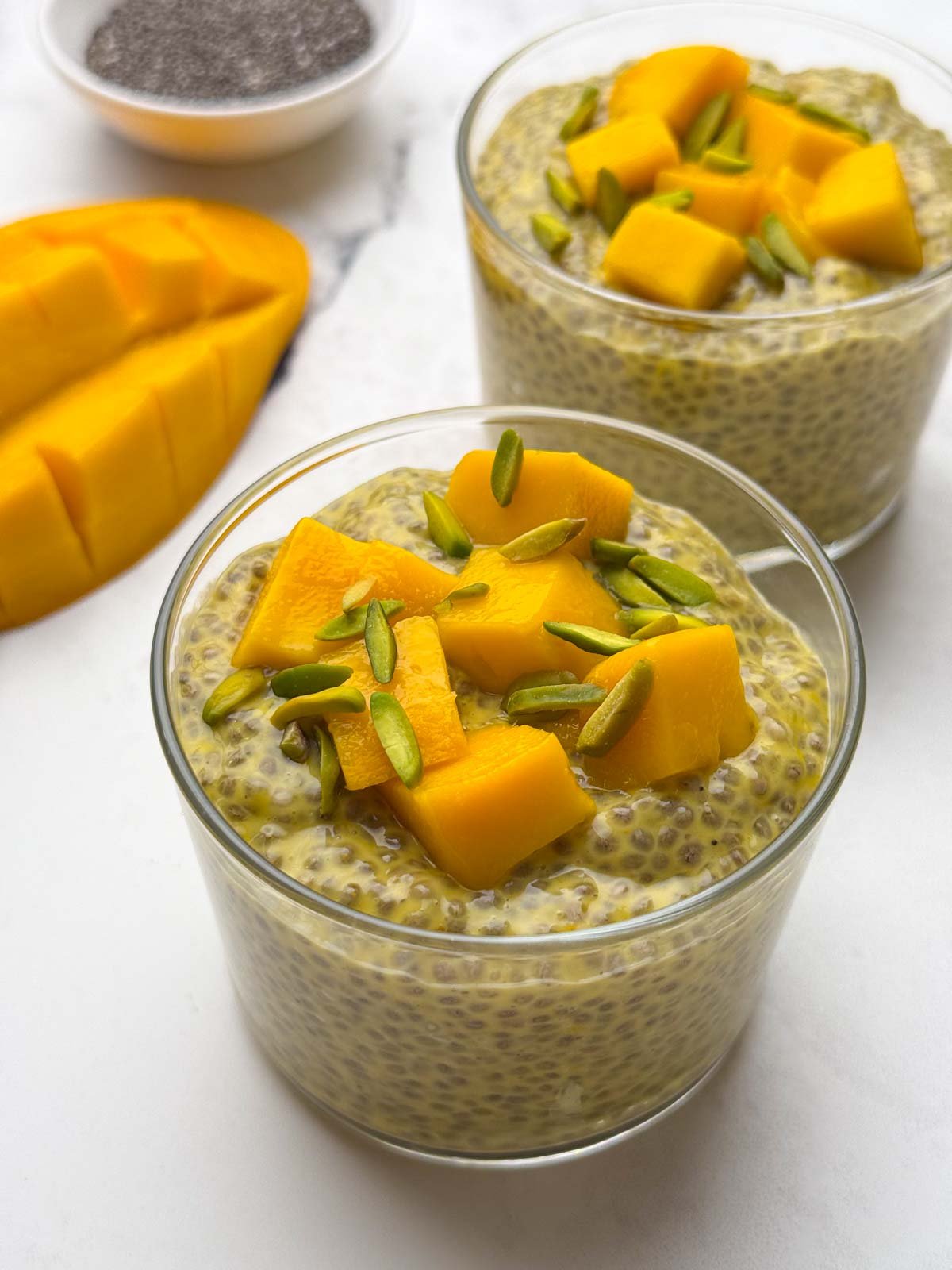 Two glass bowls of mango coconut milk chia pudding topped with diced mango and pistachios along with mango and chia seeds on the side.