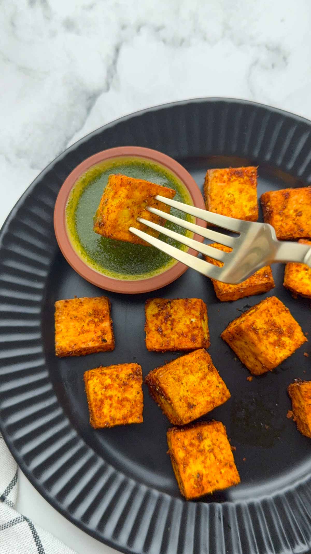 Air fried crispy paneer cubes on the black plate and using a golden fork dipping 1 piece of paneer in green chutney.