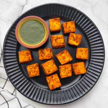 Air fried crispy paneer cubes on the black plate with green mint chutney on the side.