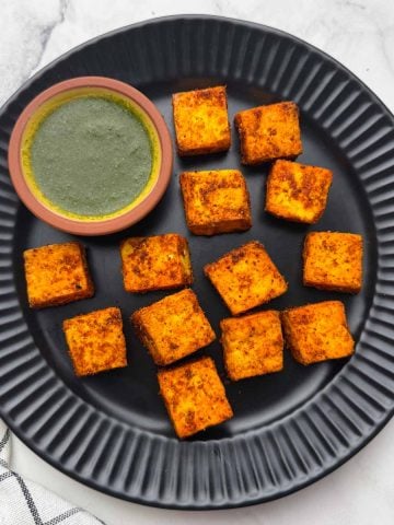Air fried crispy paneer cubes on the black plate with green mint chutney on the side.