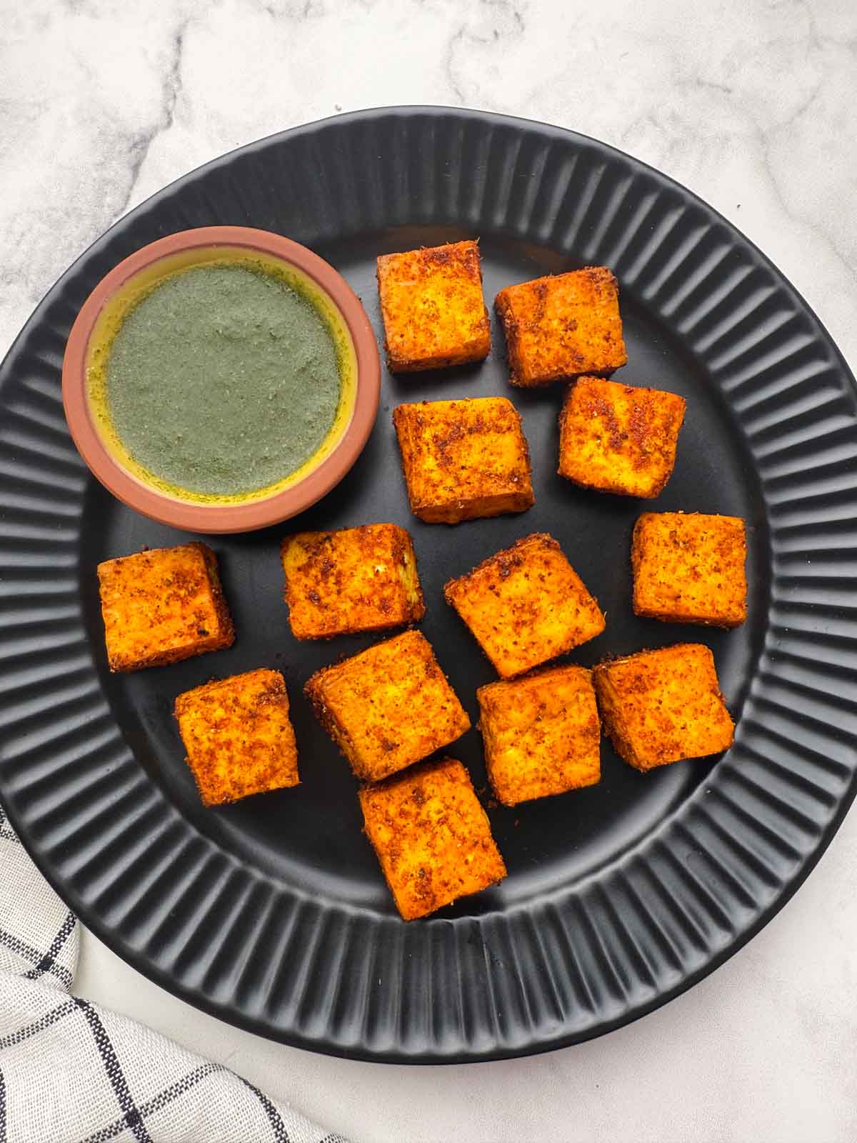 Air fried crispy paneer cubes on the black plate with green mint chutney on the side.