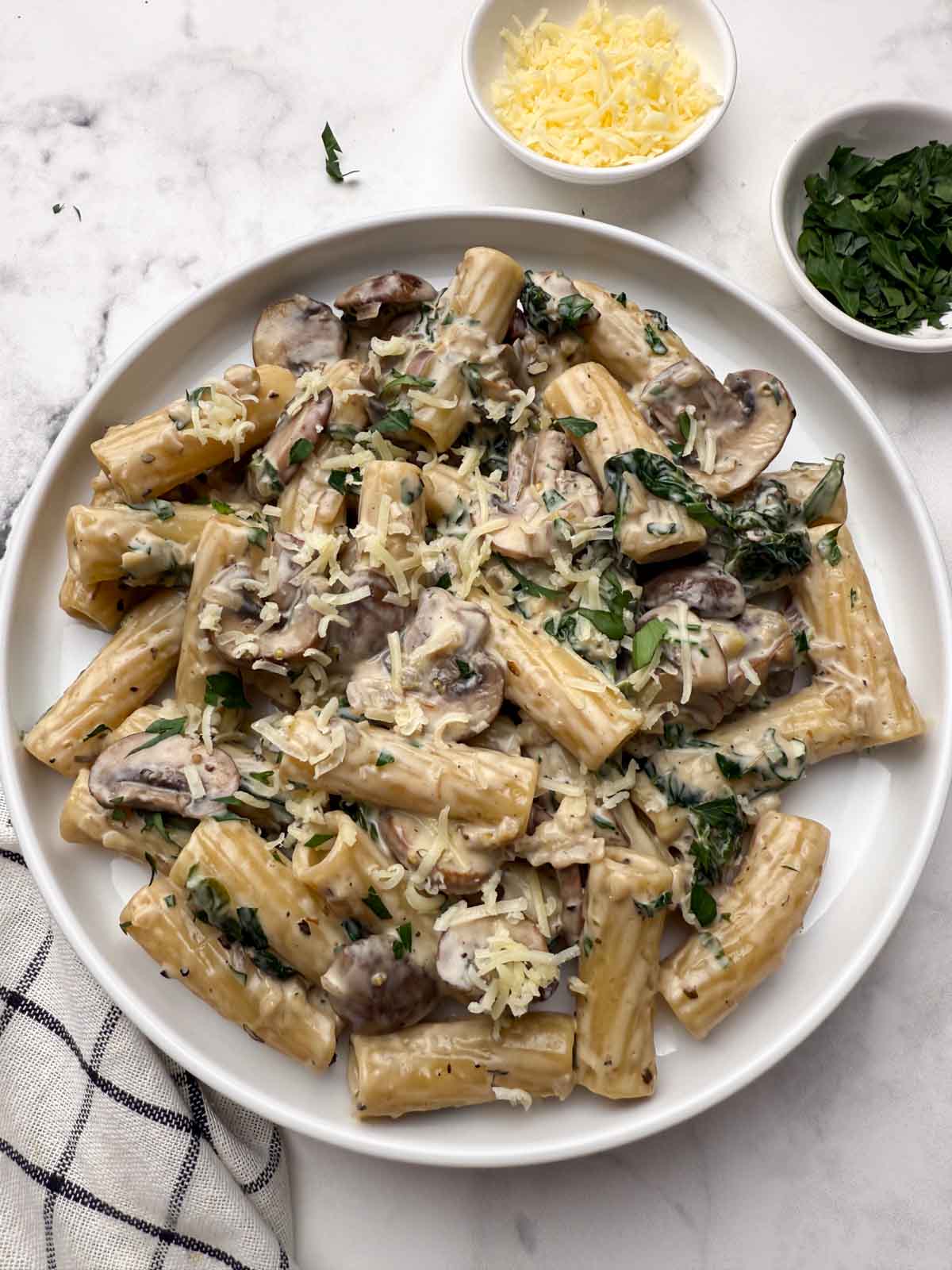 Creamy mushroom spinach pasta on a white plate with a side of cheese and fresh parsley in bowls.