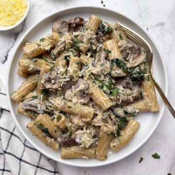Creamy mushroom pasta served on a white plate with a spoon on it, along with a side of cheese and parsley in bowls.