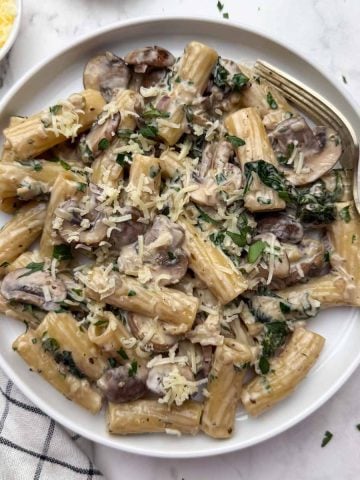 Creamy mushroom pasta served on a white plate with a spoon on it, along with a side of cheese and parsley in bowls.
