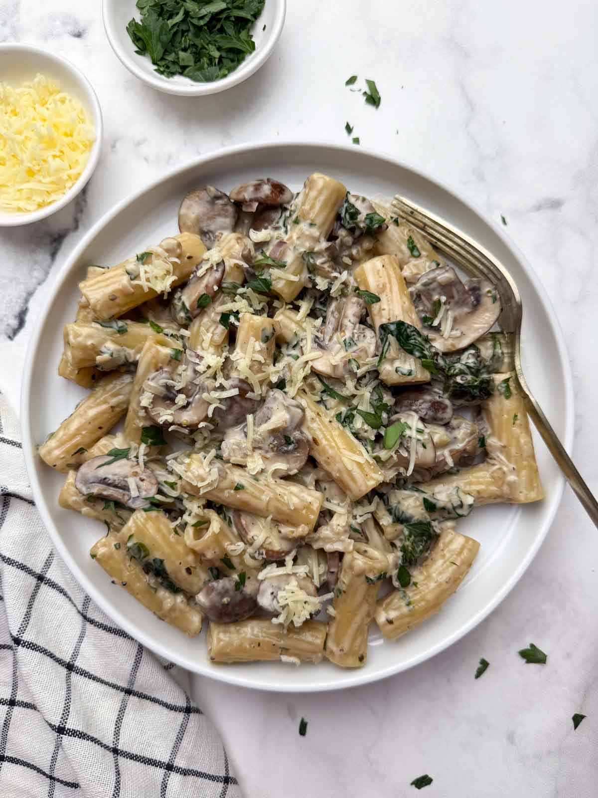 Creamy mushroom pasta served on a white plate with a spoon on it, along with a side of cheese and parsley in bowls.
