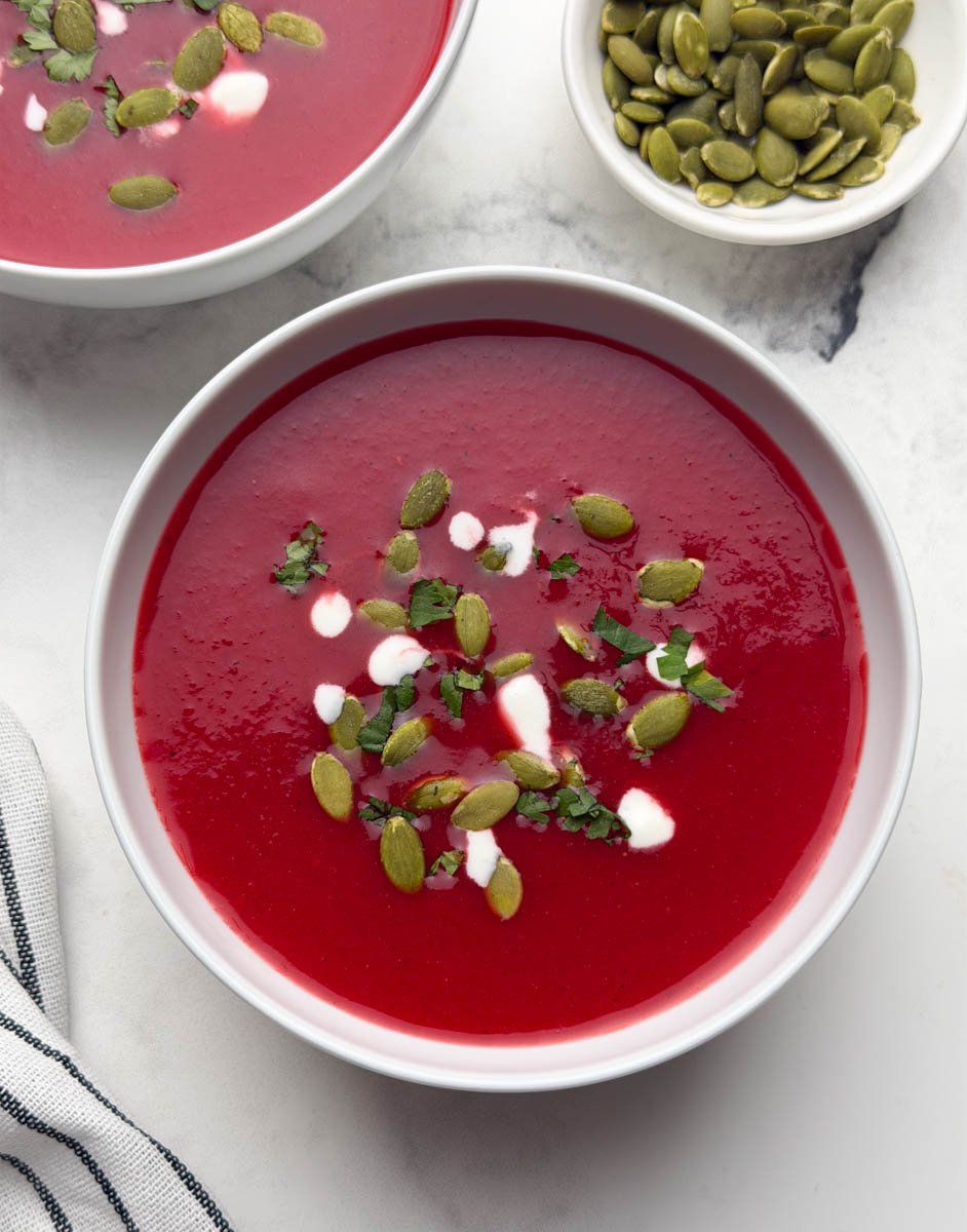 Two bowls of beetroot tomato soup garnished with pumpkin seeds, coriander leaves and cream with a spoon and also a small bowl of pumpkin seeds on the side.