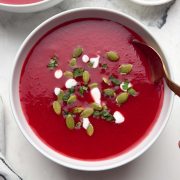 Two bowl of beetroot tomato soup garnished with pumpkin seeds, coriander leaves and cream with a spoon and also a small bowl of pumpkin seeds on the side.