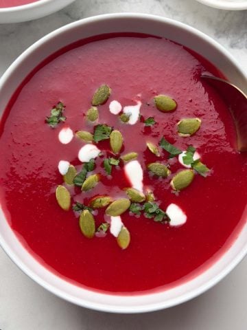 Two bowl of beetroot tomato soup garnished with pumpkin seeds, coriander leaves and cream with a spoon and also a small bowl of pumpkin seeds on the side.