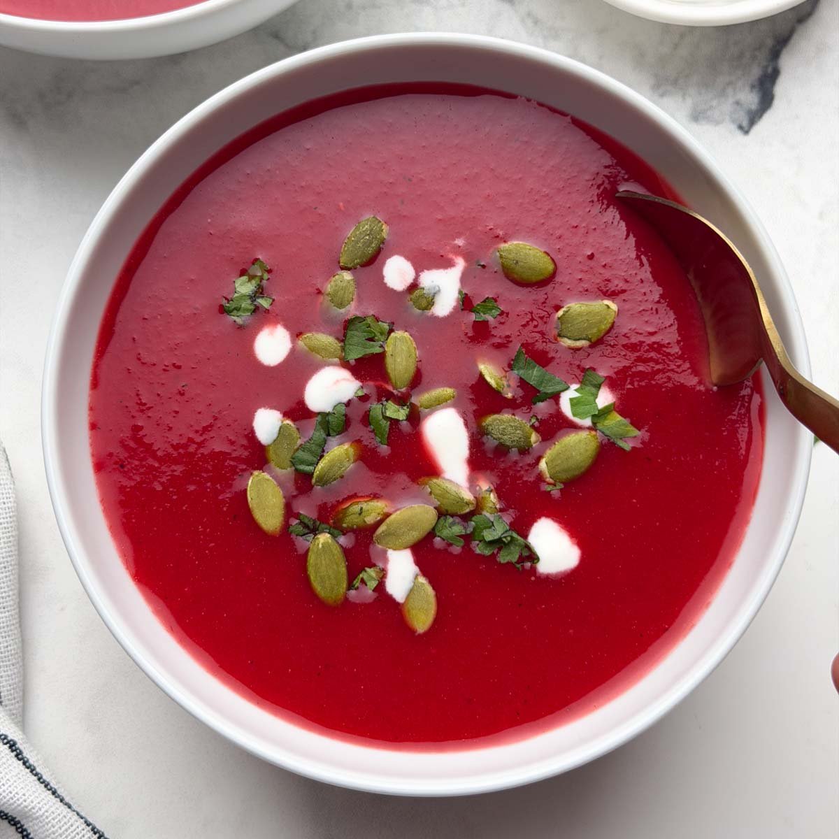 Two bowl of beetroot tomato soup garnished with pumpkin seeds, coriander leaves and cream with a spoon and also a small bowl of pumpkin seeds on the side.