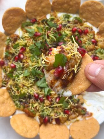 Indian papdi chaat dip served on a platter with papdi arranged, and one papdi being held with a scoop of the dip. Pomegranate, coriander and sev on the side and marigold garland on the side for decoration.