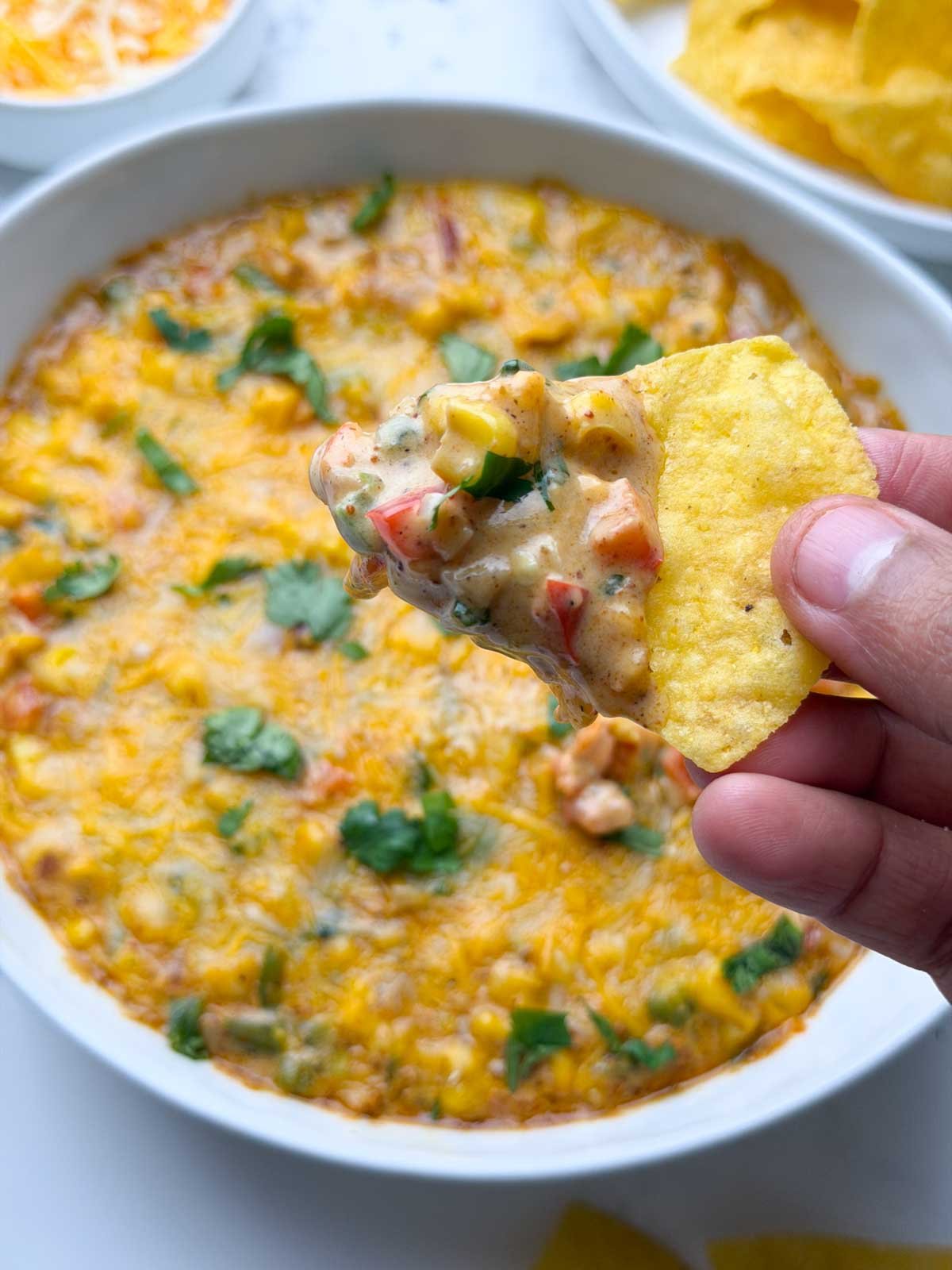 Baked masala corn dip served in a white bowl, with tortilla chips full of dip holding in the hand and tortilla chips on the side.