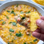 Baked masala corn dip served in a white bowl, with tortilla chips full of dip holding in the hand and tortilla chips on the side.
