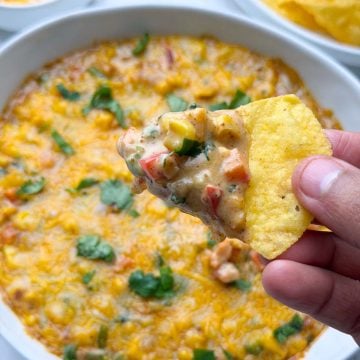 Baked masala corn dip served in a white bowl, with tortilla chips full of dip holding in the hand and tortilla chips on the side.