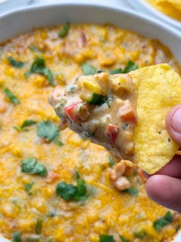 Baked masala corn dip served in a white bowl, with tortilla chips full of dip holding in the hand and tortilla chips on the side.