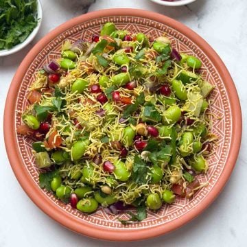 Edamame chaat served on a brown plate, garnished with sev. Small bowls of chopped coriander, sev, and pomegranate are placed alongside.