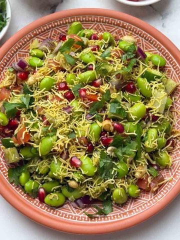 Edamame chaat served on a brown plate, garnished with sev. Small bowls of chopped coriander, sev, and pomegranate are placed alongside.