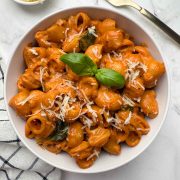 A bowl of creamy tomato pasta garnished with basil leaf, also garlic bread, cheese and a golden fork kept on the side.