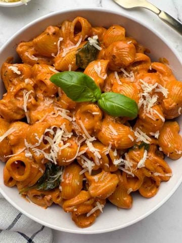 A bowl of creamy tomato pasta garnished with basil leaf, also garlic bread, cheese and a golden fork kept on the side.